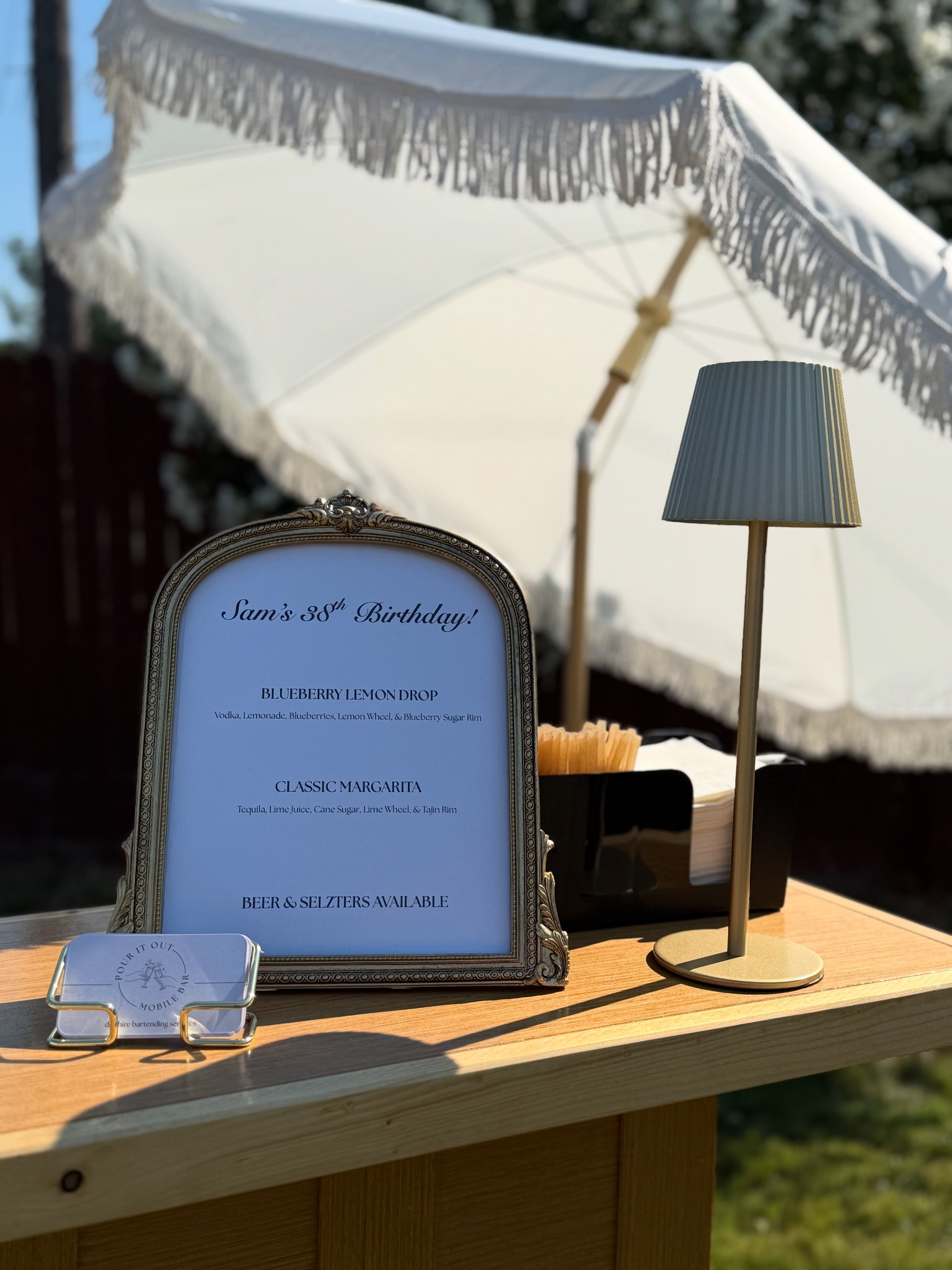Custom cocktail menu card in an ornate framed sign for a birthday celebration, with a brass table lamp on the bar