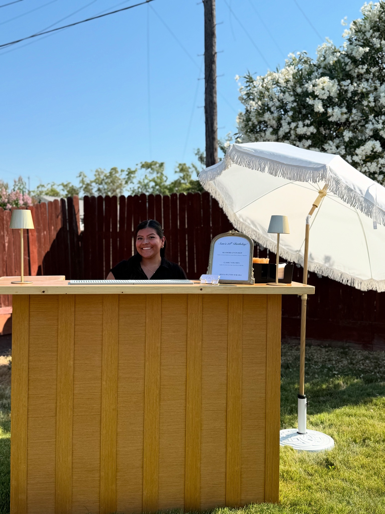 Pour It Out bartender smiling behind the mobile bar at an outdoor birthday party under a fringed umbrella