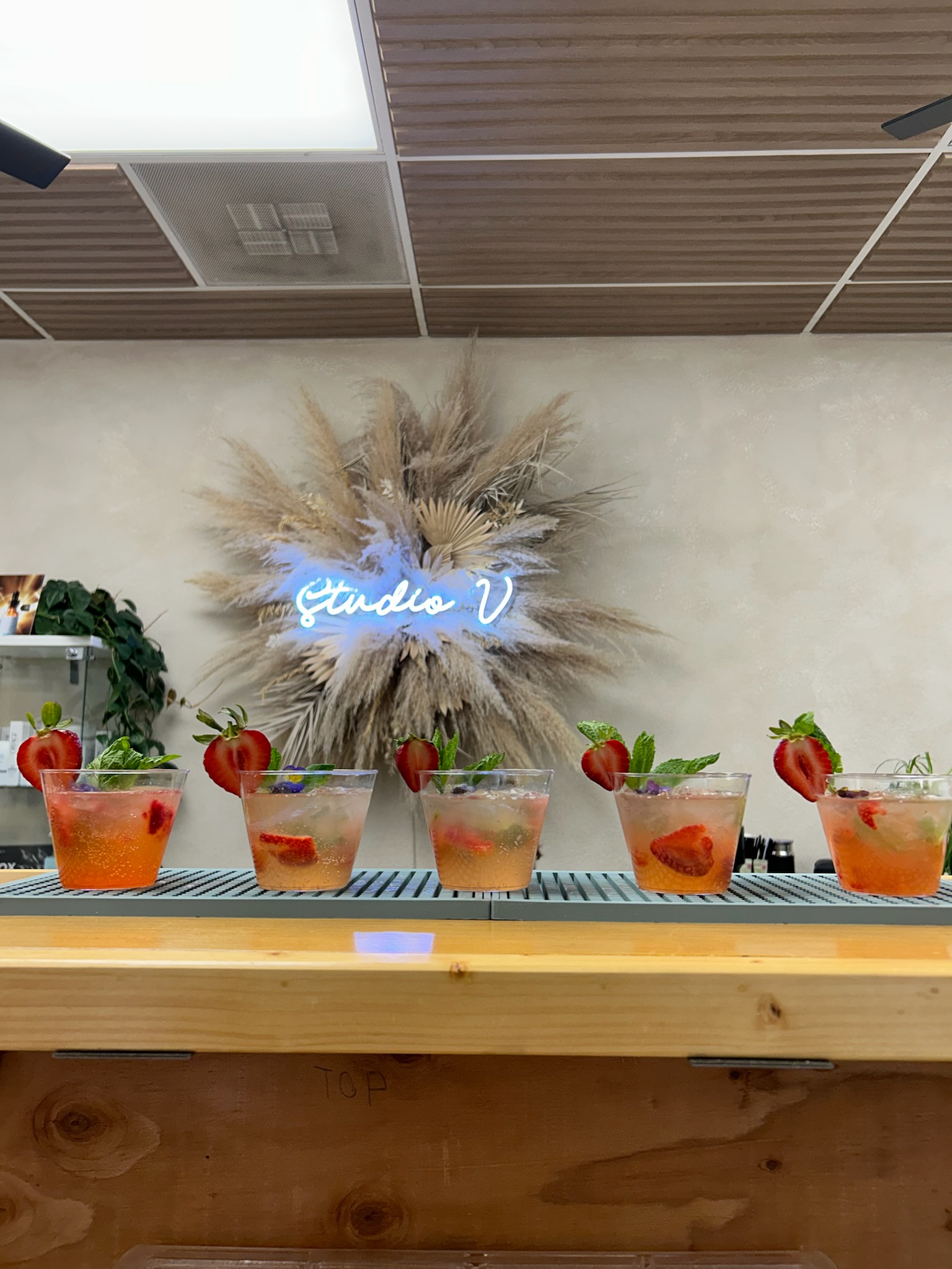 Five strawberry garden cocktails lined up on the bar in front of a Studio V neon sign with a pampas grass backdrop