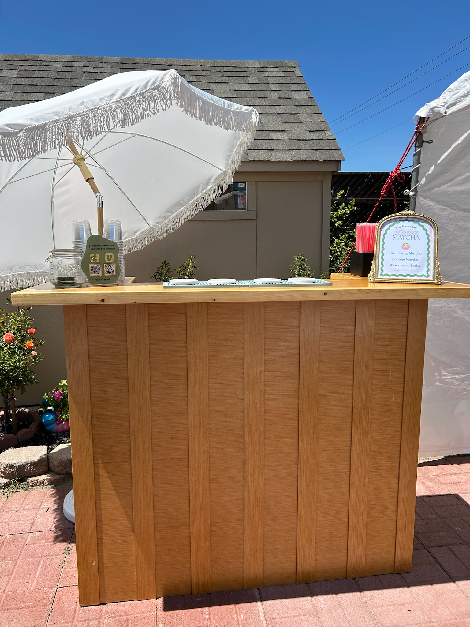 Outdoor bar top detail with a framed birthday cocktail menu and brass table lamp under a fringed umbrella