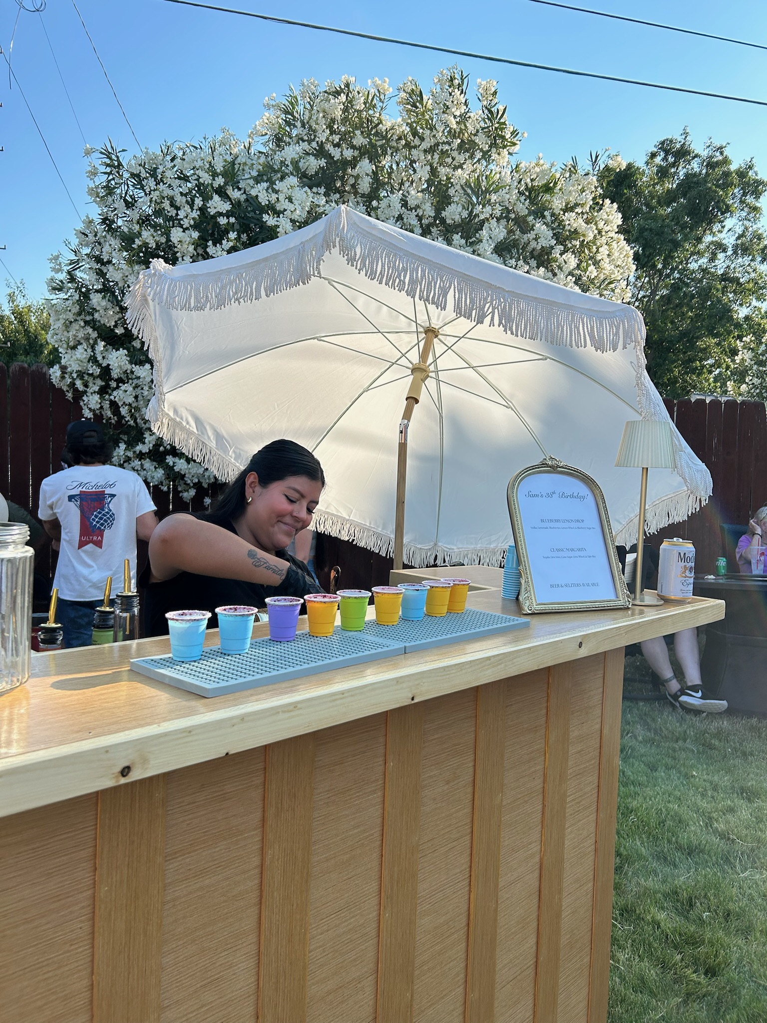 Owner smiling behind the mobile bar with a row of colorful margarita cups lined up under the fringed umbrella