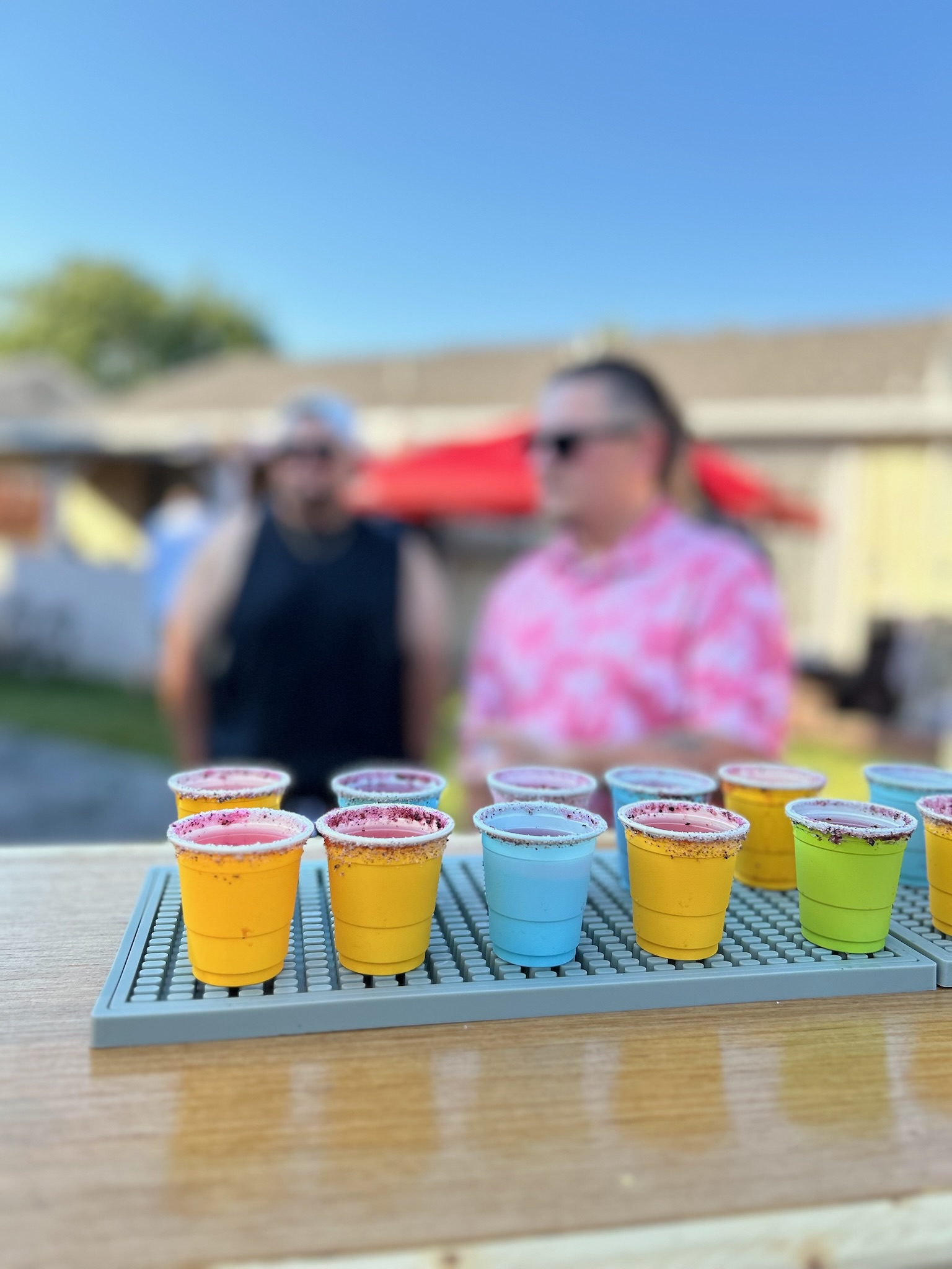 Colorful yellow, blue, and green margarita cups lined up on the bar mat at a lively backyard birthday party