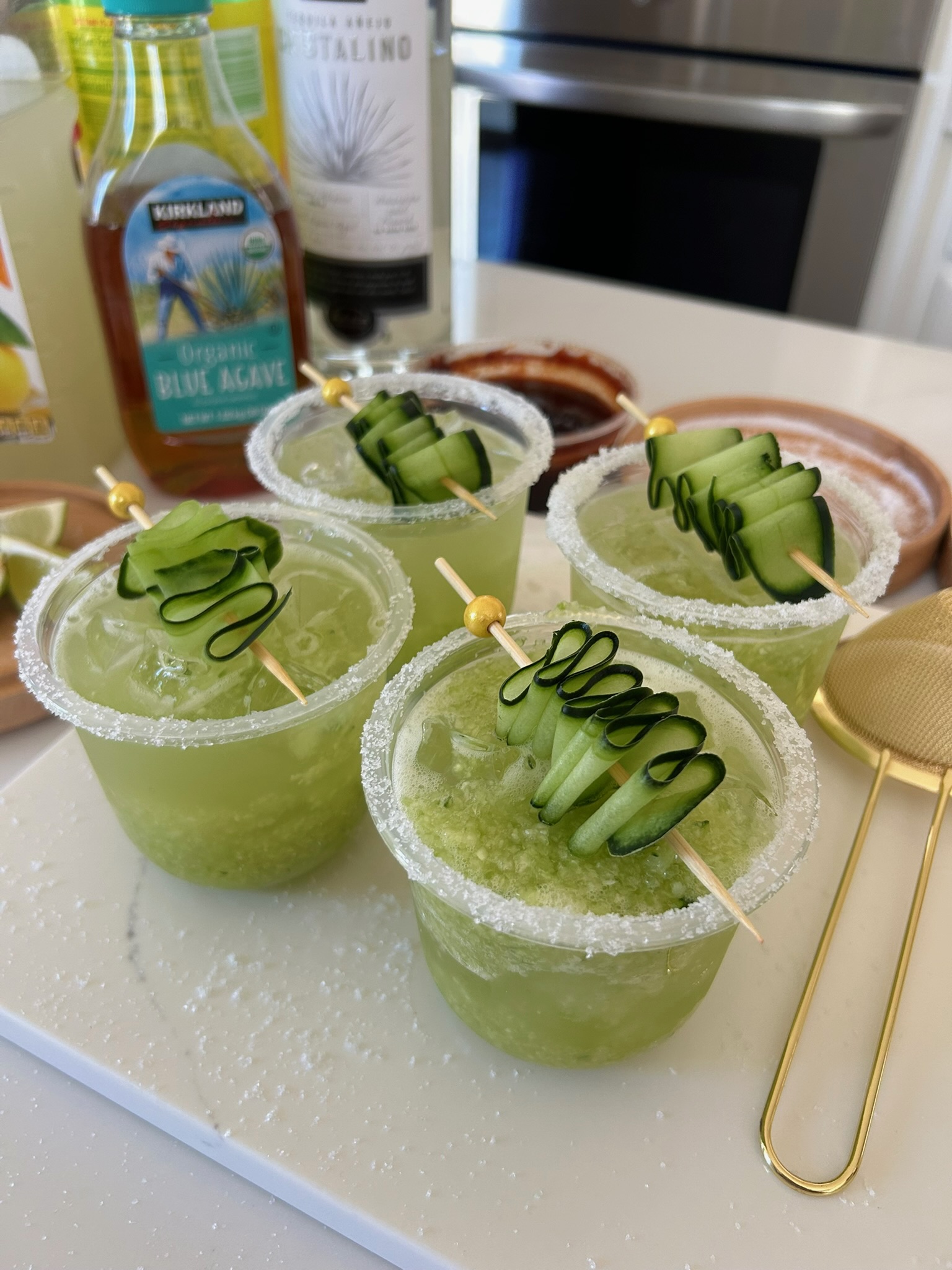 Four cucumber jalapeño margaritas with salted rims and spiraled cucumber garnish lined up on a white marble counter
