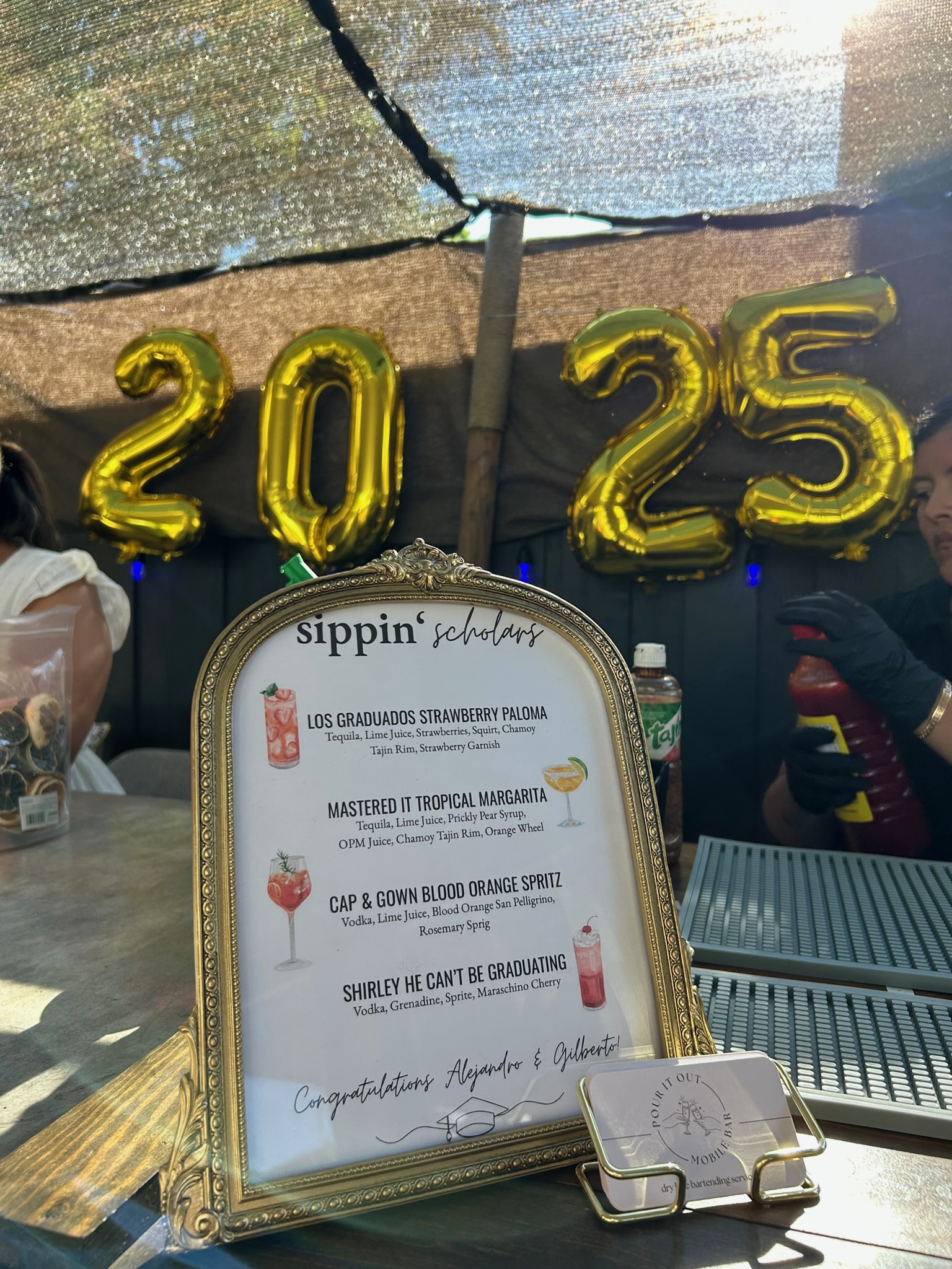 Sippin' Scholars custom graduation cocktail menu in a gold frame surrounded by 2025 gold balloon decorations