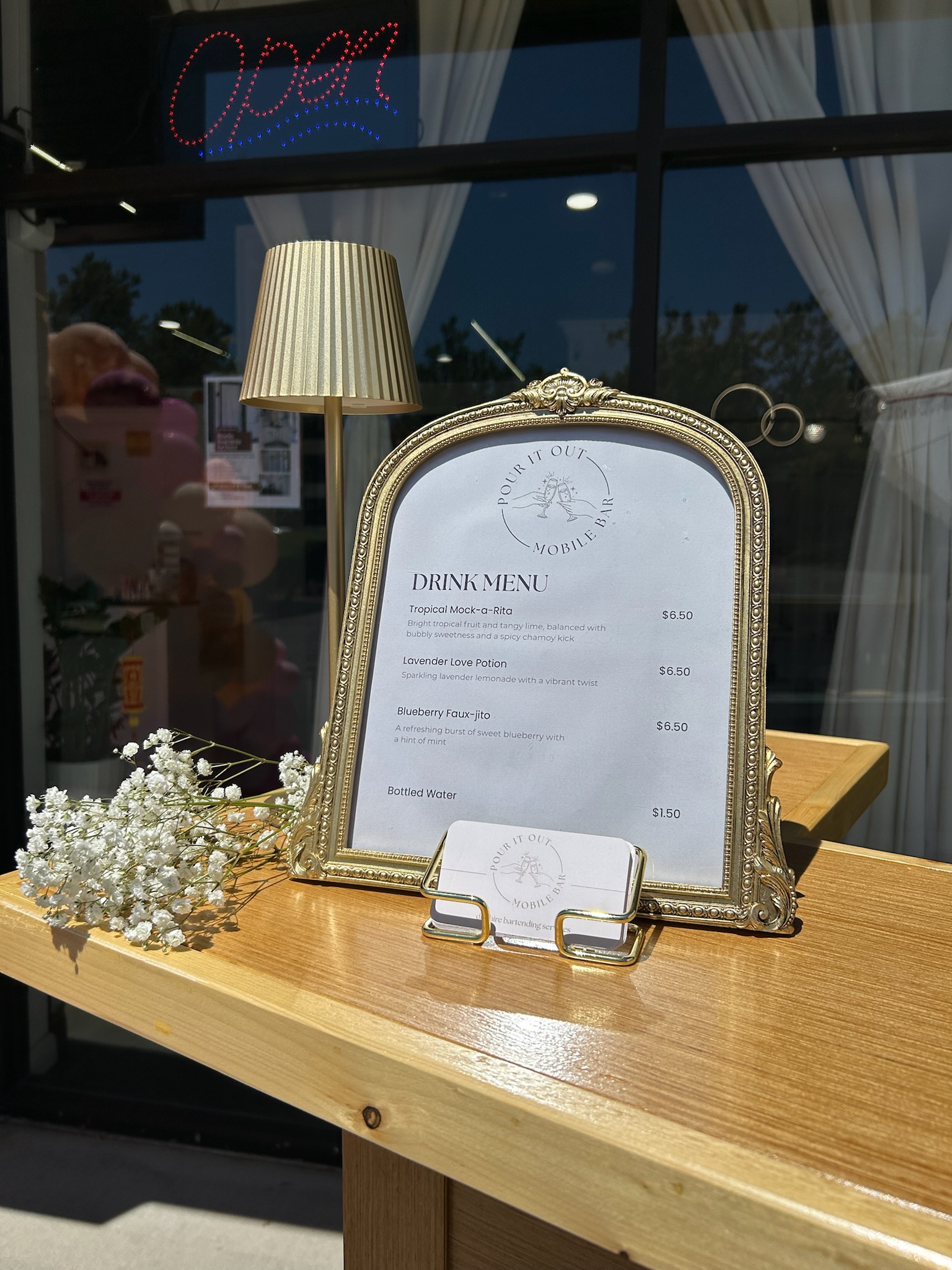 Pour It Out drink menu displayed in a gold arched frame with baby's breath flowers and business cards on the bar top