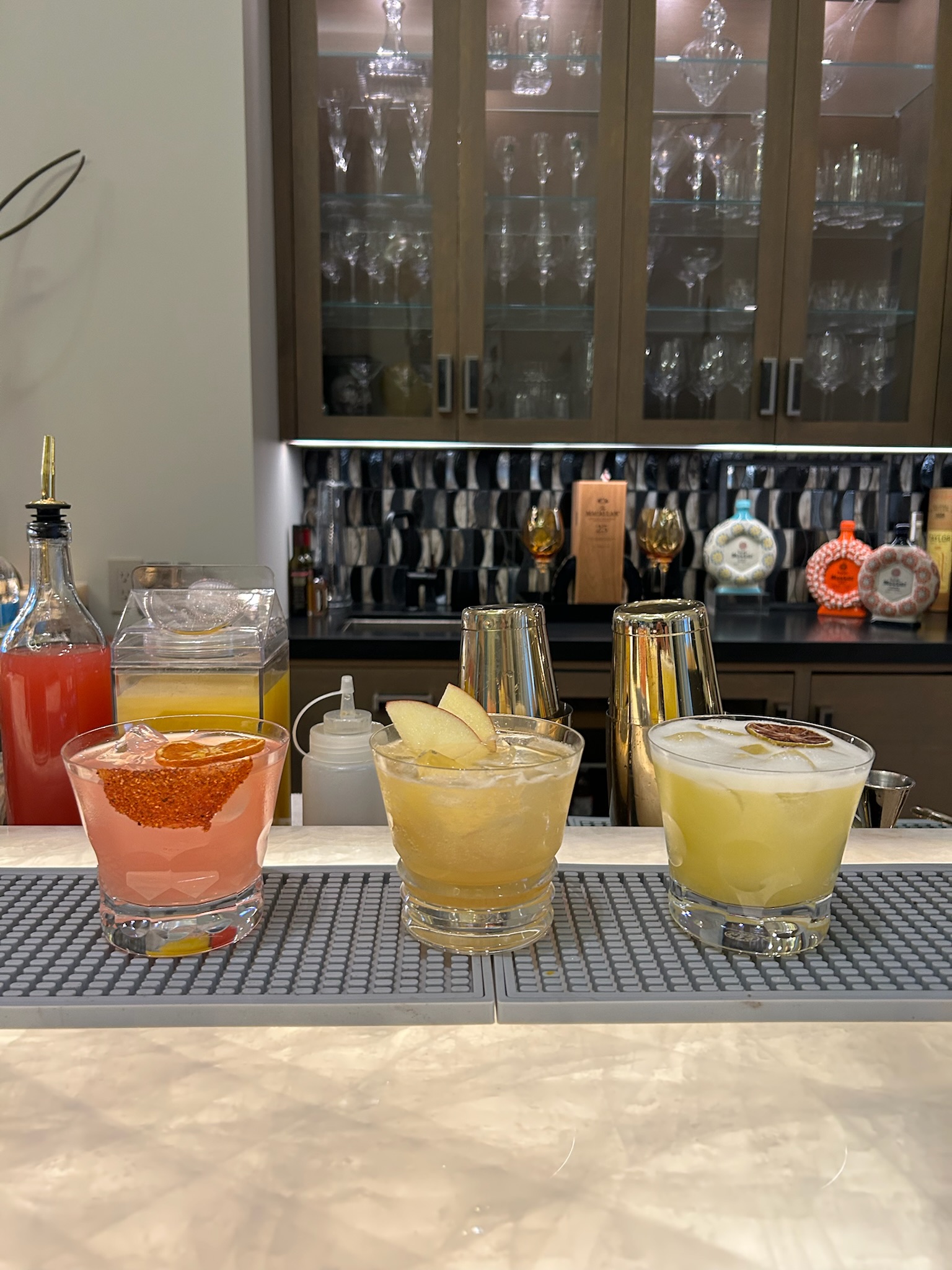 Three craft cocktails: a pink Paloma, a gold apple sour, and a creamy citrus sour, lined up on the bar at an elegant home event