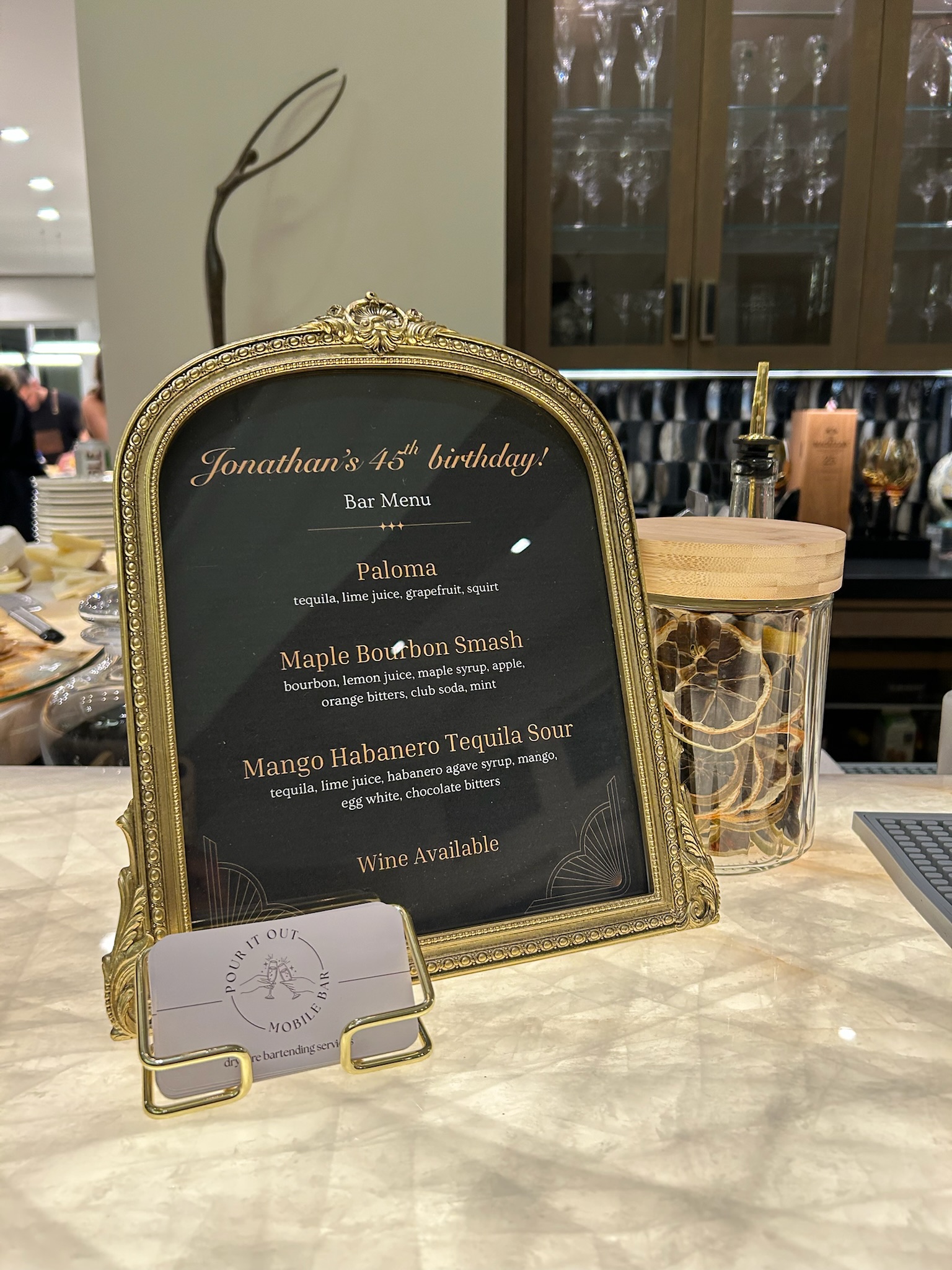 Jonathan's birthday cocktail menu in an ornate gold frame beside a gold cocktail shaker on an elegant marble bar top