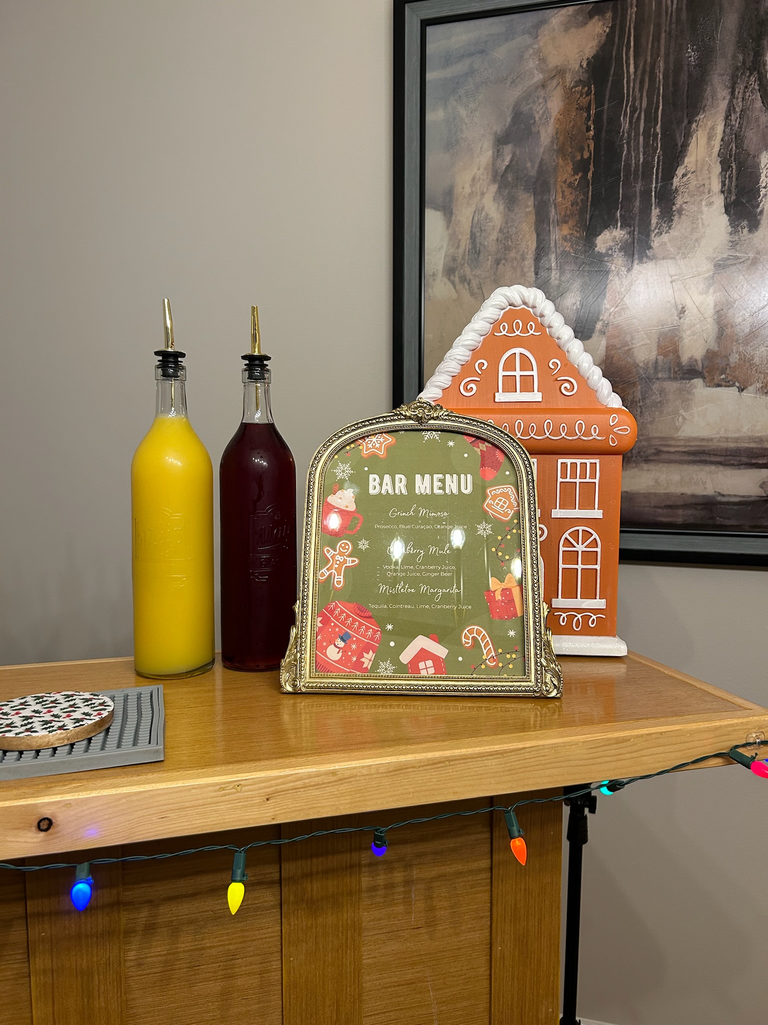 Christmas holiday bar menu in a gold frame with a gingerbread house decoration and bottles of fresh-squeezed juice wrapped in festive lights